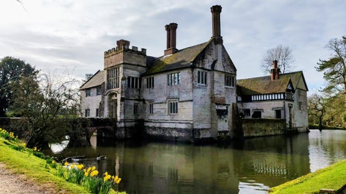 The south-west corner of the house at Baddesley Clinton, Warwickshire, seen across the moat with spring daffodils at the edge